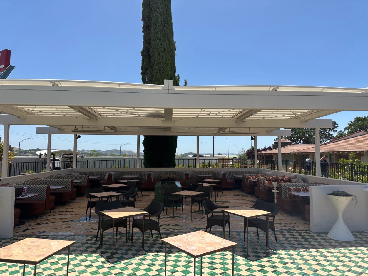 Outdoor patio with tables and chairs, surrounded by greenery and blue sky.
