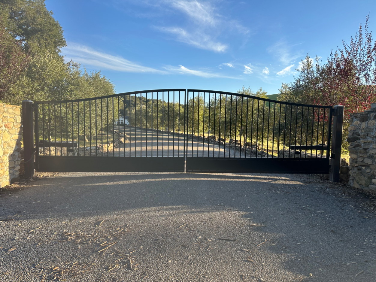 Black wrought iron gate at the entrance of a gravel driveway surrounded by trees.