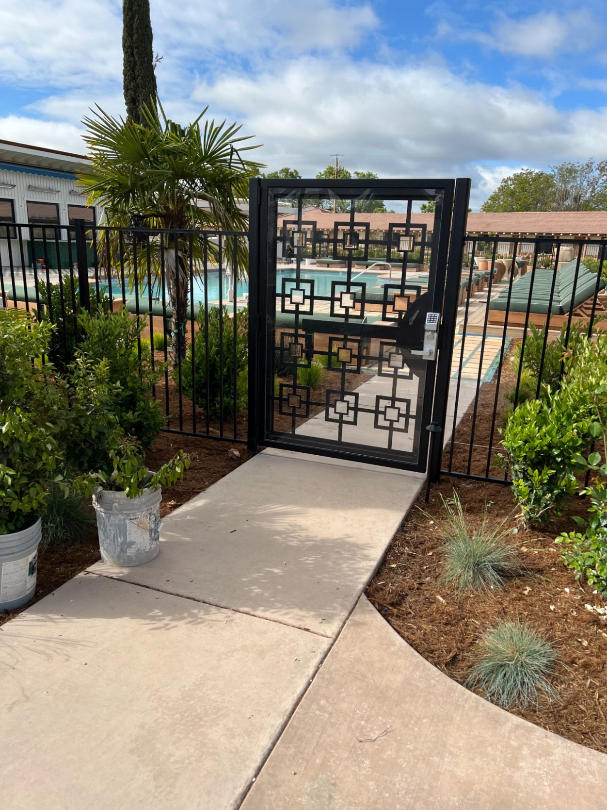 Black gate leading to a pool area, surrounded by lush greenery and decorative planters.