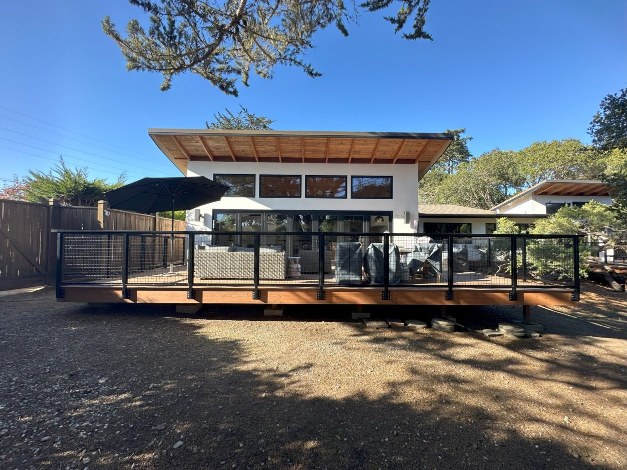 Modern house with a wooden deck, large windows, and a patio umbrella surrounded by greenery.